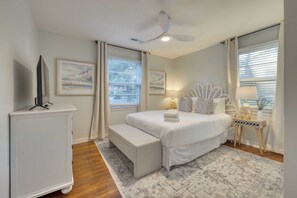 Une chambre à coucher comprenant un lit, un banc, un ventilateur de plafond, des œuvres d’art et une fenêtre avec des rideaux.