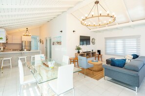 A modern kitchen with a stainless steel refrigerator, a dining area with a glass table, and a living room with a grey sofa and a television.