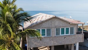 A two-story building with a stone facade and a flat roof, featuring a covered patio area and a view of the ocean.