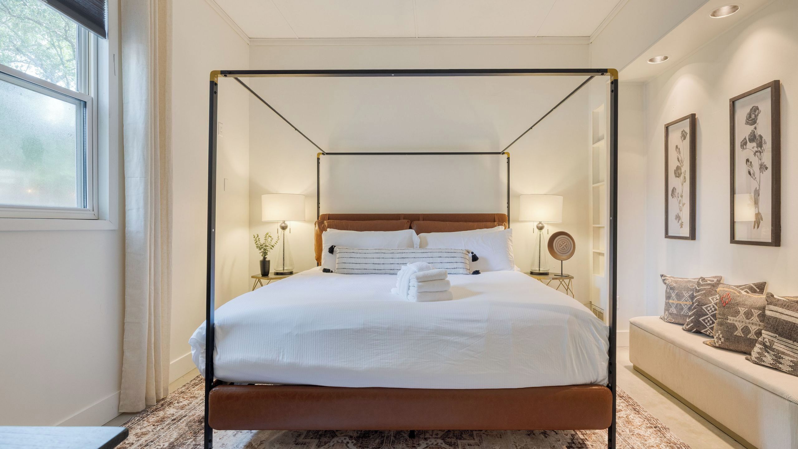 Una cama con dosel con sábanas blancas, una mesita de noche con lámpara y un ventilador de pared.