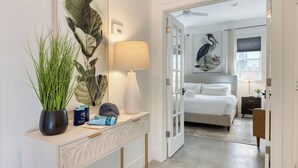 A bedroom with a bed, a nightstand, a lamp, a large plant, and a painting on the wall. - The Cannon Cottage by Avantstay Old Mt. Pleasant Gem Near Sullivans Island (Mount Pleasant)