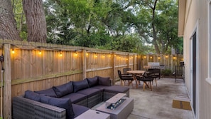 Un patio al aire libre con cerramiento de madera, luces colgantes, una parrilla, un sofá y una mesa de comedor.