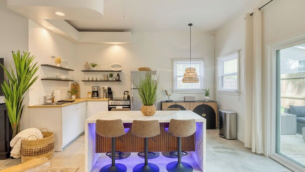 A modern kitchen with a central island, built-in shelves, and a dining area with bar stools. - The Cannon Cottage by Avantstay Old Mt. Pleasant Gem Near Sullivans Island (Mount Pleasant)