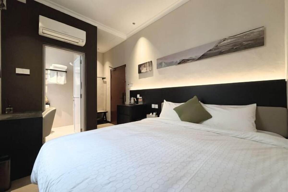 A hotel room with a large bed, a bedside table, a chair, an air conditioning unit, a door, and a wall-mounted artwork.