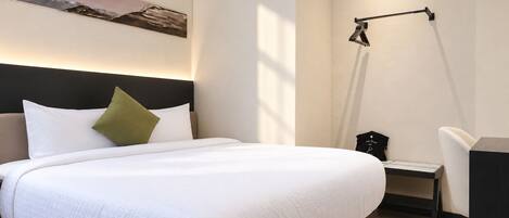 A neatly made bed with white bedding and a single green pillow, a desk with a keyboard and a chair, and a floor lamp against the wall.