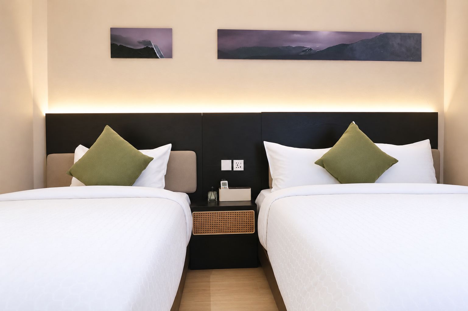 A hotel room with two beds, each with white bedding and green pillows, a nightstand with a lamp, and two framed pictures on the wall.