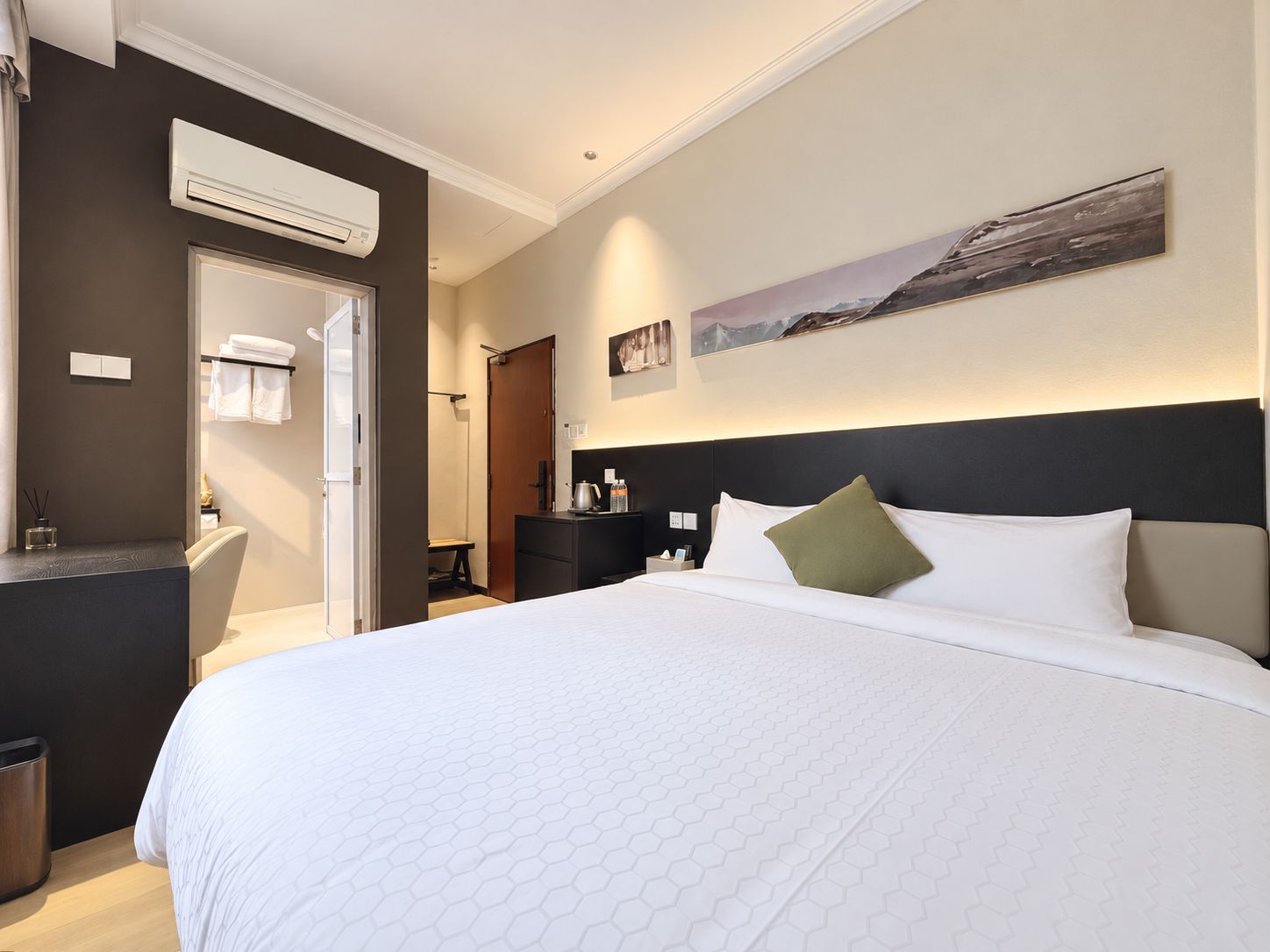 Habitación de hotel con una cama grande, una mesita de noche, un equipo de aire acondicionado, un cuadro en la pared y una puerta que da al baño.