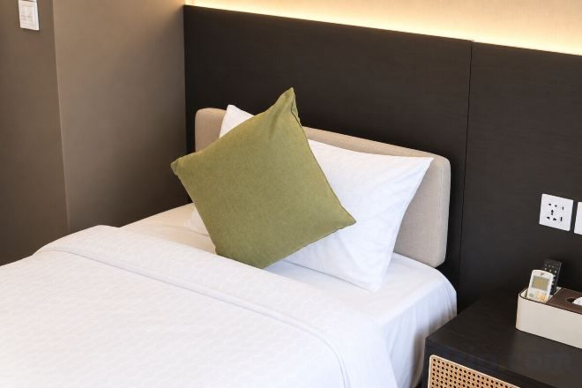 A neatly made bed with white bedding and a green pillow, a bedside table with a lamp, and a power outlet on the wall.