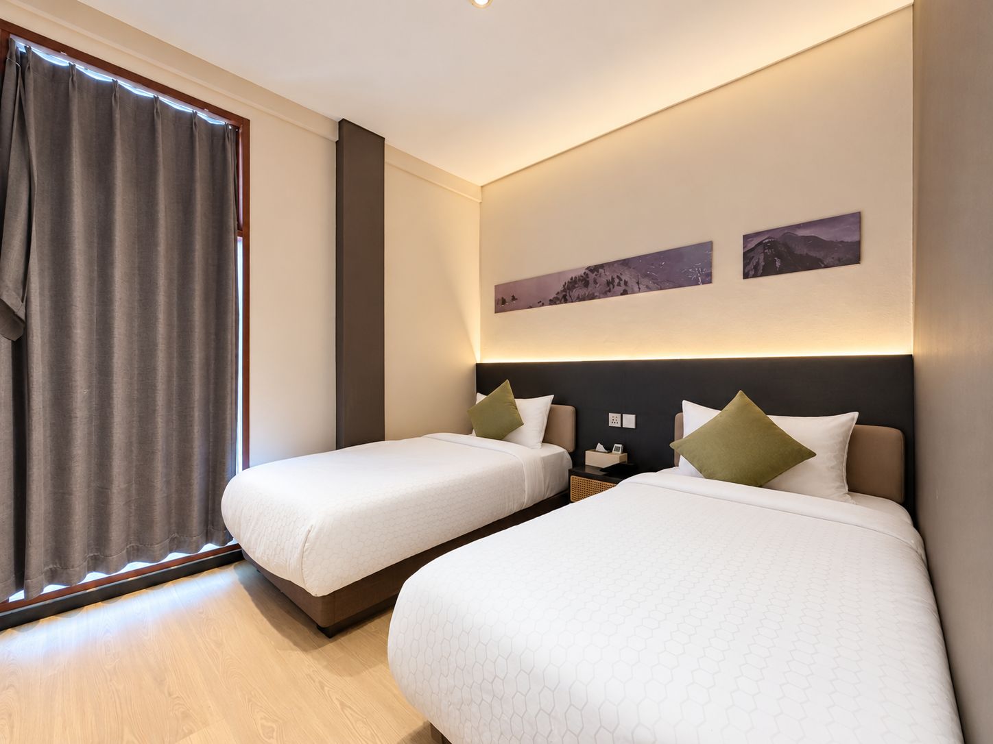 Quarto de hotel com duas camas, uma janela grande com cortinas, mesa de cabeceira e uma obra de arte na parede.