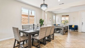 A dining area with a dark wooden table and chairs, a chandelier, and large windows with a view of a brick building. - Casa Campo 5BR Desert Oasis w/ Pool, Firepit, & Outdoor Lounge (Indio)
