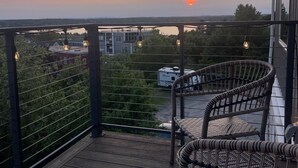 Una terraza de madera con sillas de mimbre, vista a un atardecer y una caravana al fondo.