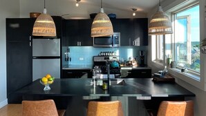 Una cocina moderna con una isla central, tres lámparas colgantes y vistas de la ciudad a través de amplios ventanales.