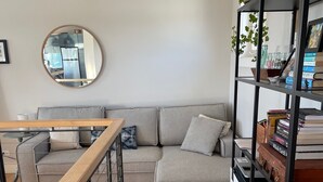 Una sala moderna con un sofá seccional gris, un espejo redondo, una mesa de comedor de madera y una estantería negra llena de libros y objetos decorativos.