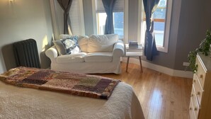 Un dormitorio con cama, sofá blanco, pequeña mesa, radiador y ventana con cortinas.