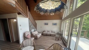Amplia sala de estar con muebles de mimbre, una escalera de piedra y ventanas grandes con vistas al exterior.