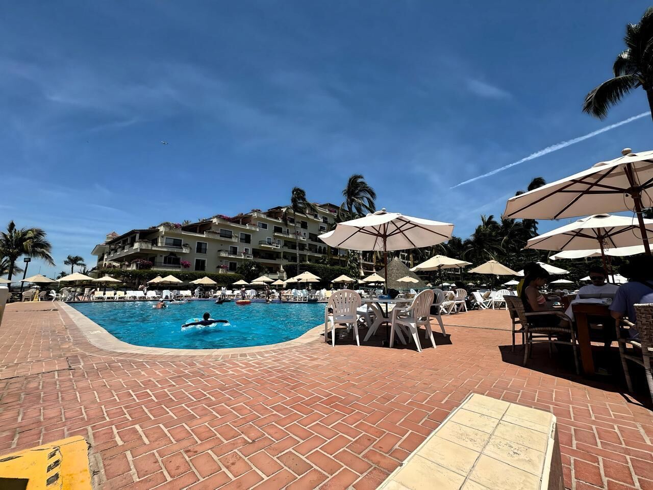 A pool area with lounge chairs and umbrellas, a brick walkway, and a building with balconies in the background.
