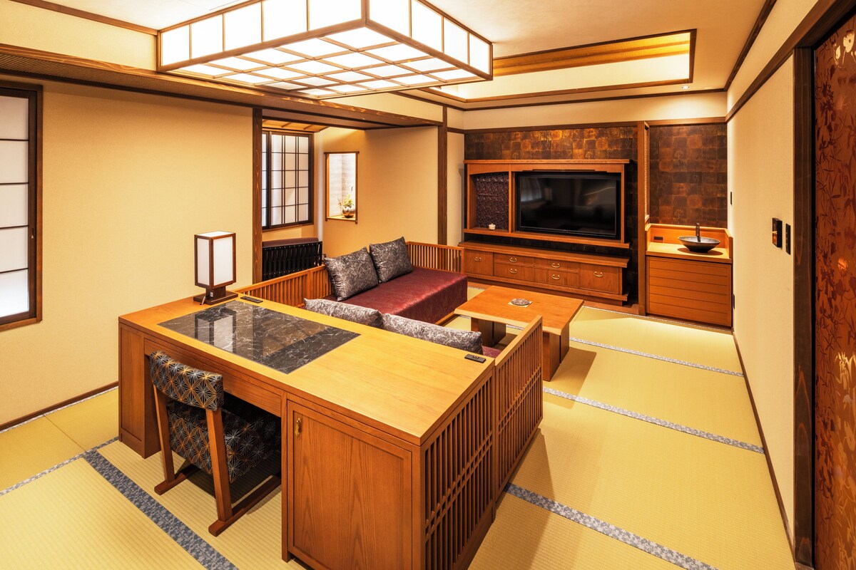 A traditional Japanese-style room with tatami flooring, a low wooden table with cushions, a television, and a small kitchen area.