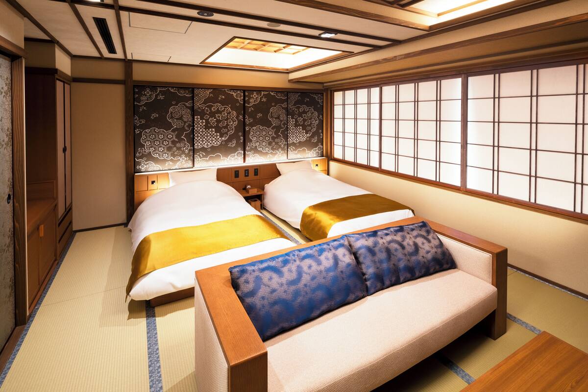 Una habitación japonesa tradicional con suelo de tatami, dos camas con ropa de cama amarilla y azul, un sofá de madera y un panel shoji.