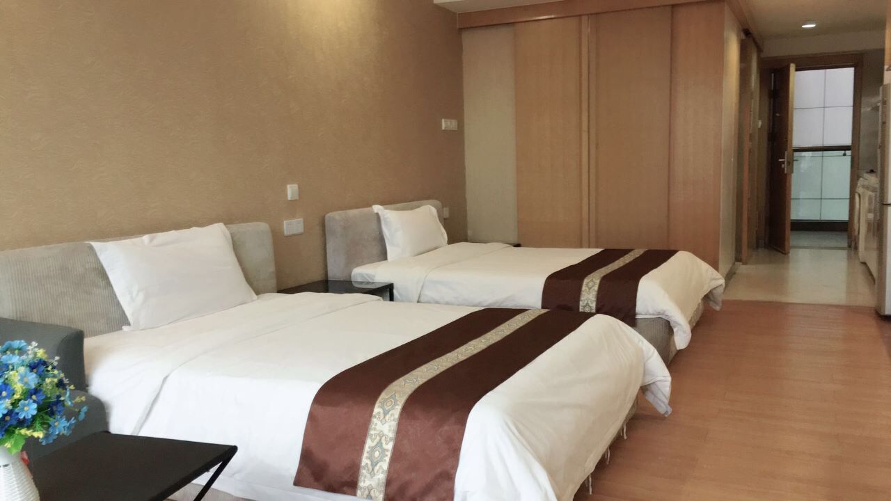 A hotel room with two beds, a small table with a vase of flowers, and a view of the hallway and balcony.