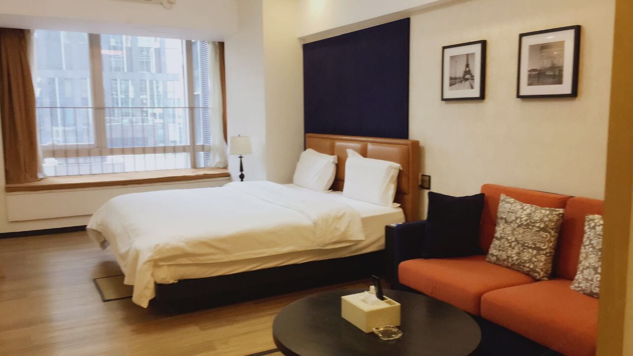 A hotel room with a bed, a sofa, a bedside table with a tissue box, and a window with blinds.