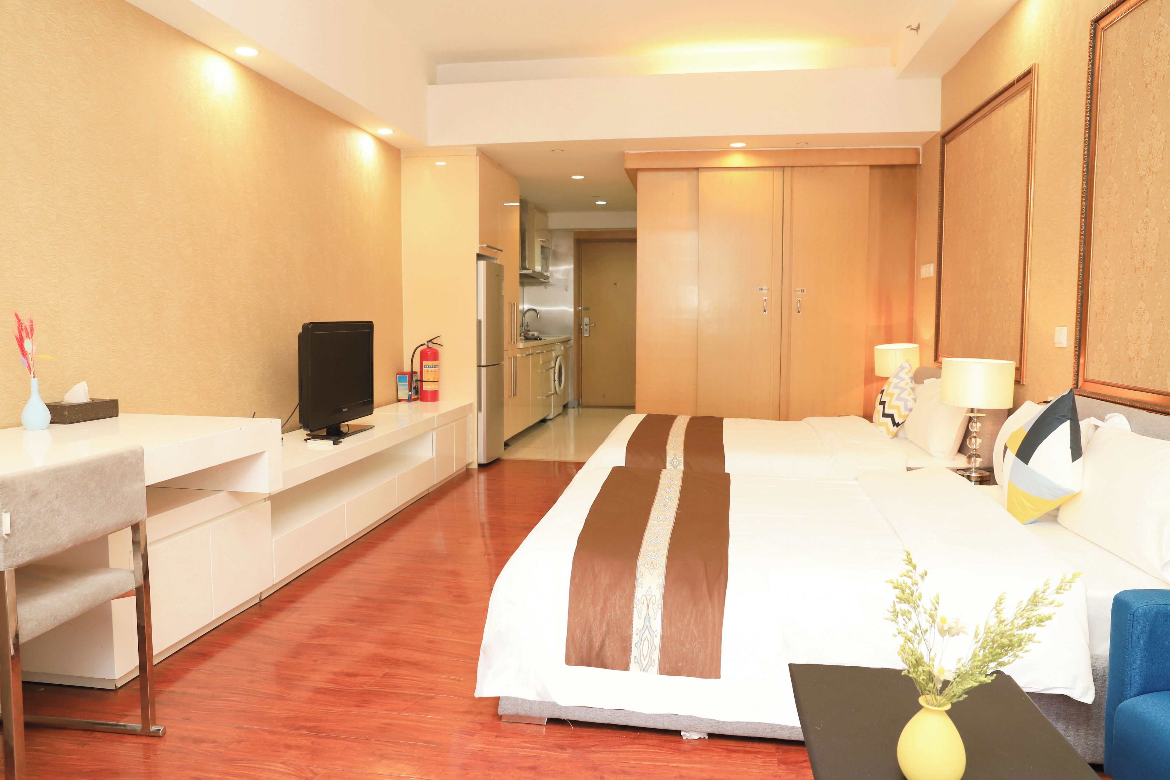 Habitación de hotel con cama, escritorio, televisión, una pequeña mesa con un florero y vistas a la cocina y al baño.