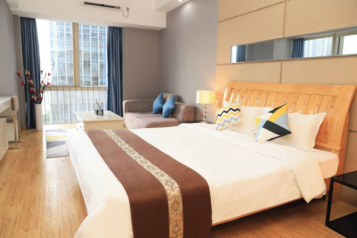 A hotel room with a large bed, a wooden headboard, a bedside table, a lamp, a chair, and a window with curtains.