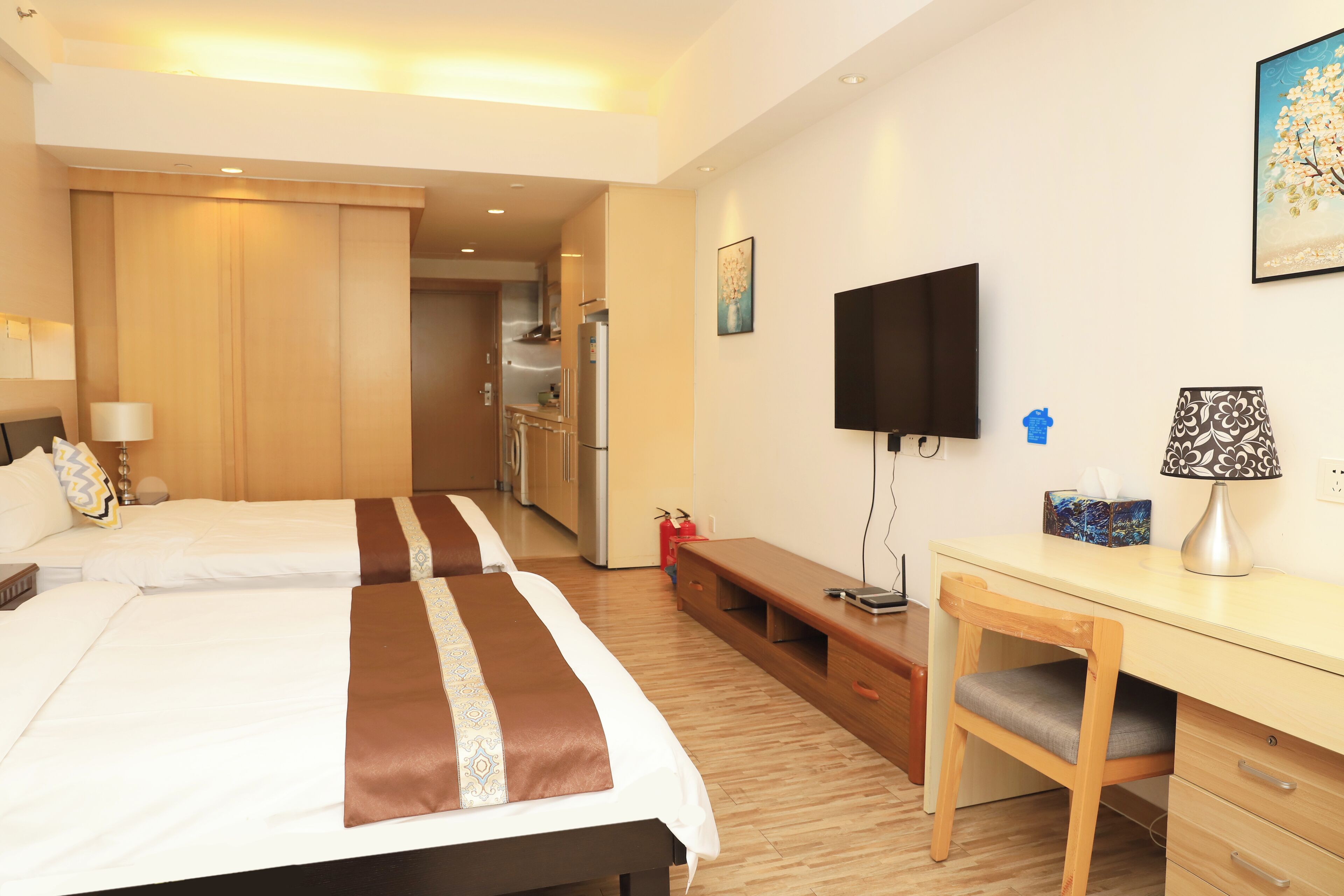 Ein Hotelzimmer mit zwei Betten, einem Schreibtisch, einem Stuhl, einem Fernseher und einer Küchenzeile im Hintergrund.