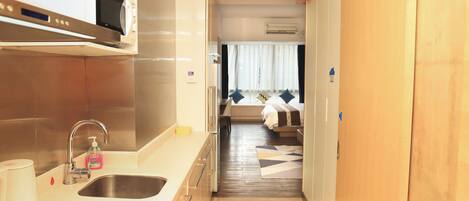 Un pasillo estrecho con una zona de cocina en un lado, una cama con ventana y cortinas en el otro, y una puerta de madera.
