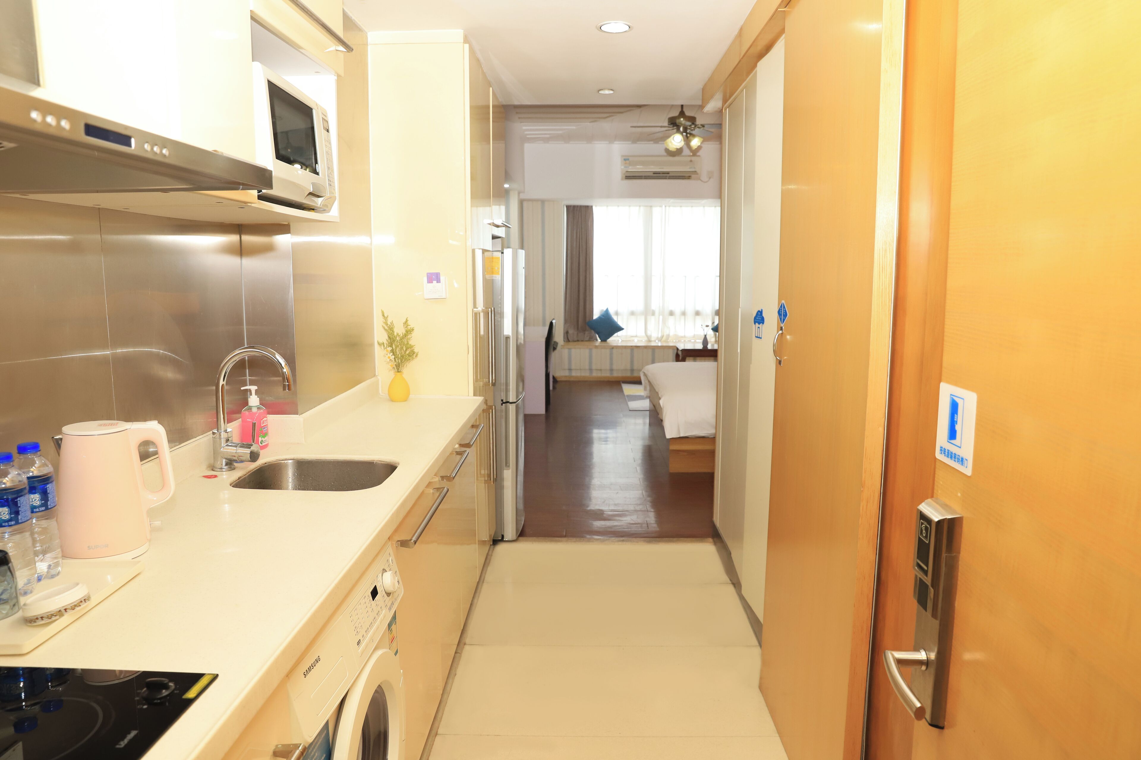 A modern kitchen with stainless steel appliances, a sink, and a microwave. There is a hallway leading to a bedroom with a bed and a window.