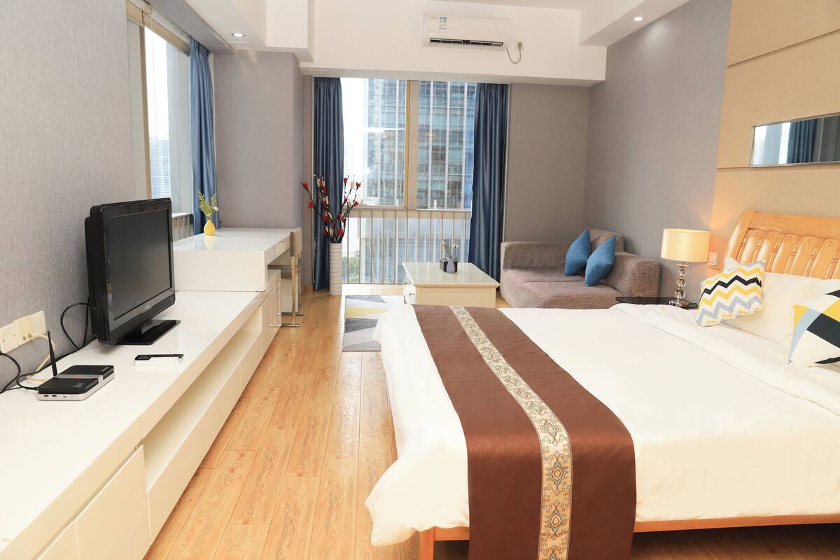 A modern hotel room with a large bed, a TV, a desk, and a balcony with a view of the city.