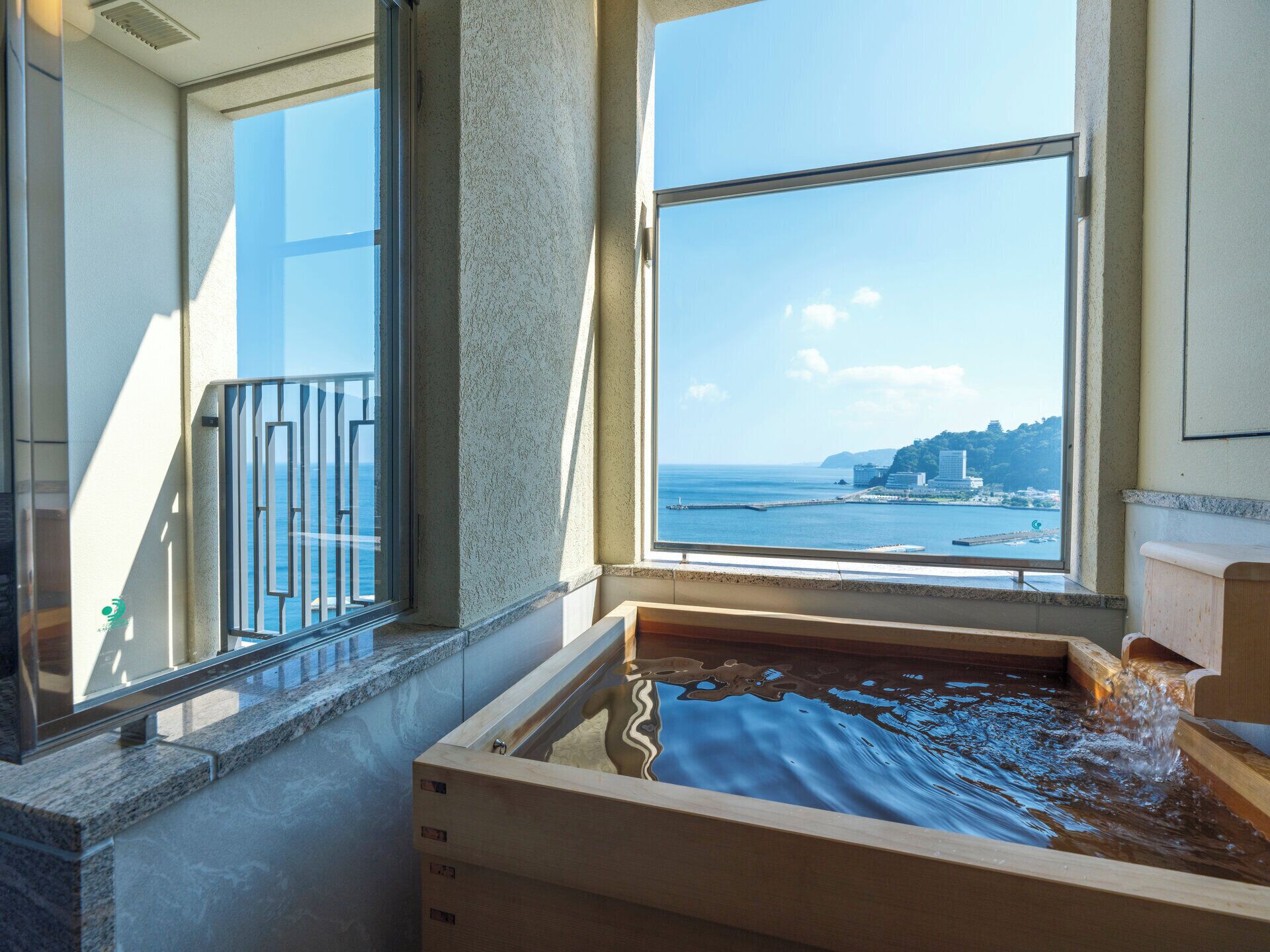 A room with a large window overlooking a coastal view, a wooden bathtub, and a balcony with a railing.