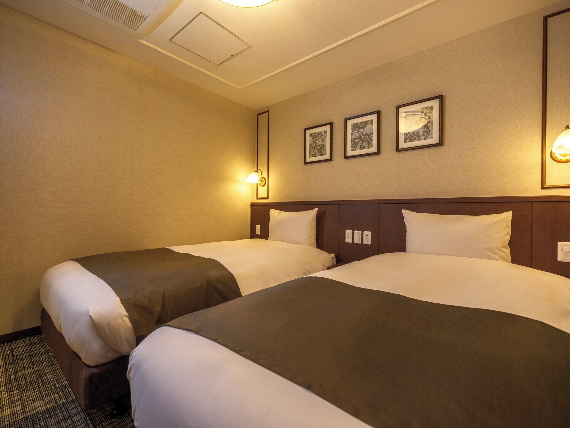 A hotel room with two beds, a headboard, a wall-mounted light, and three framed pictures.
