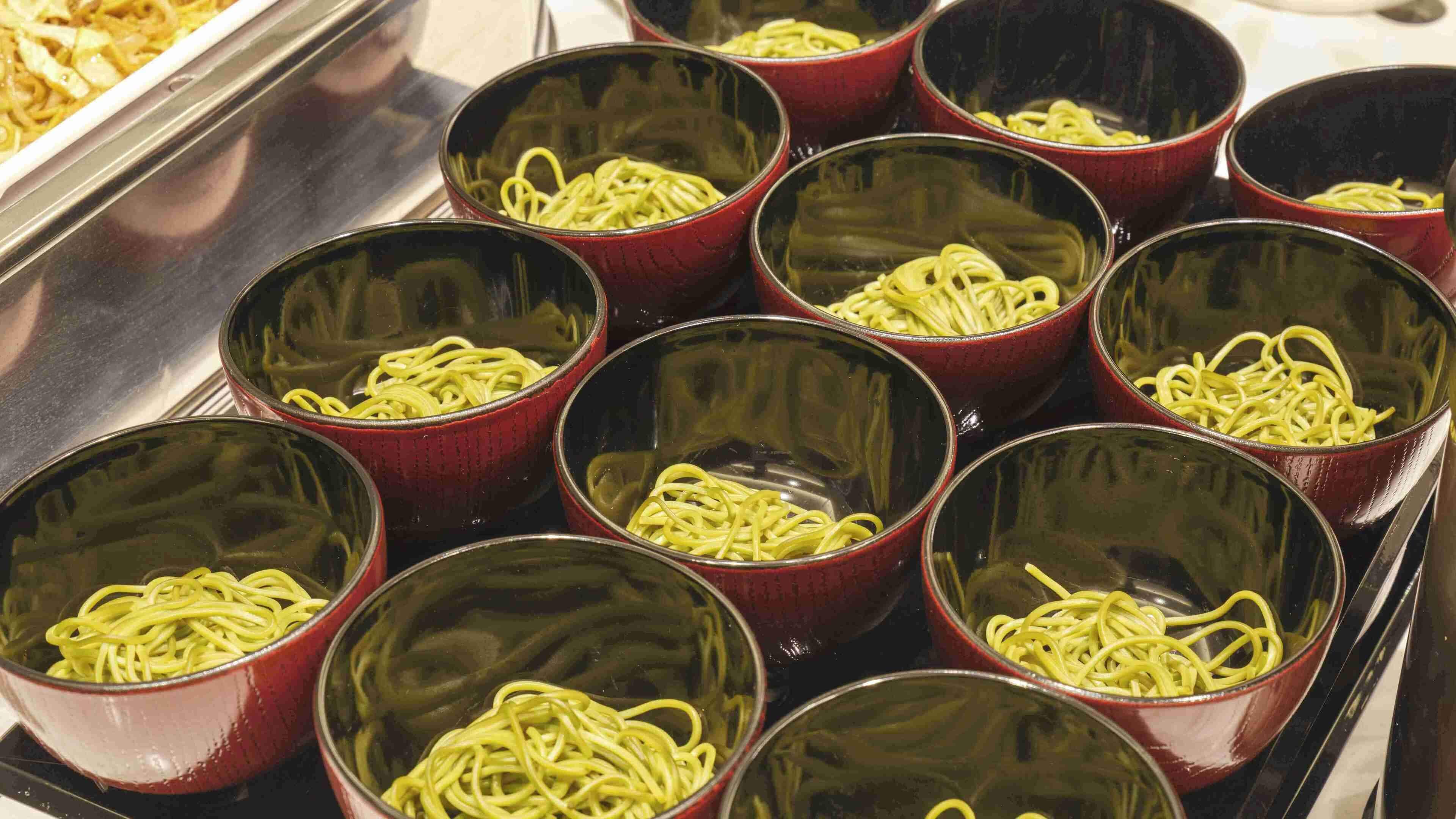 A row of red bowls containing cooked noodles on a tray.