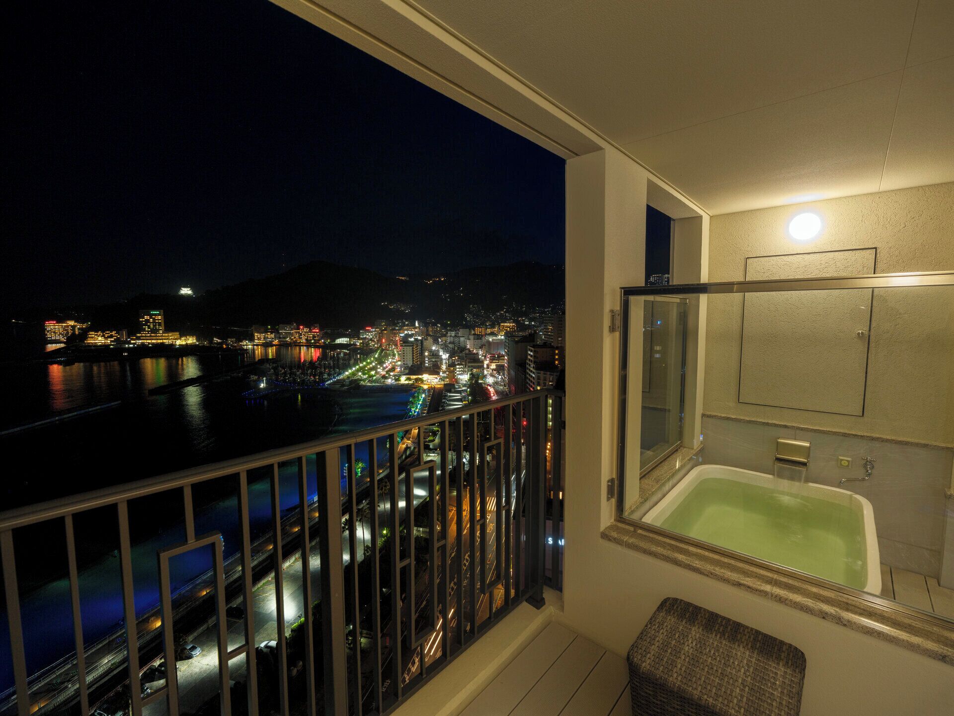 A balcony with a view of a cityscape at night, a bathtub, and a small seating area.