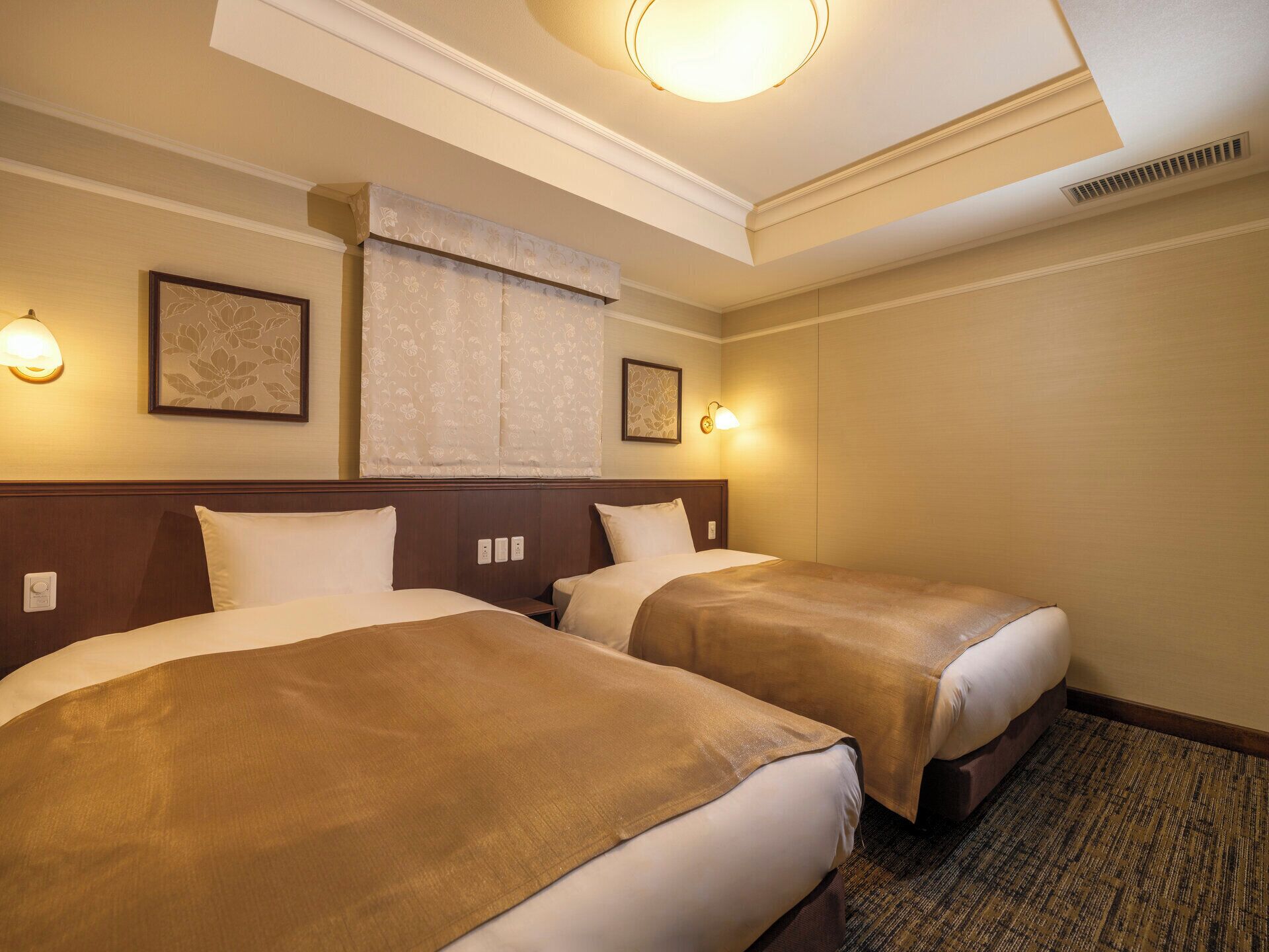 A hotel room with two beds, a large window with a curtain, wall-mounted lamps, and a ceiling light.