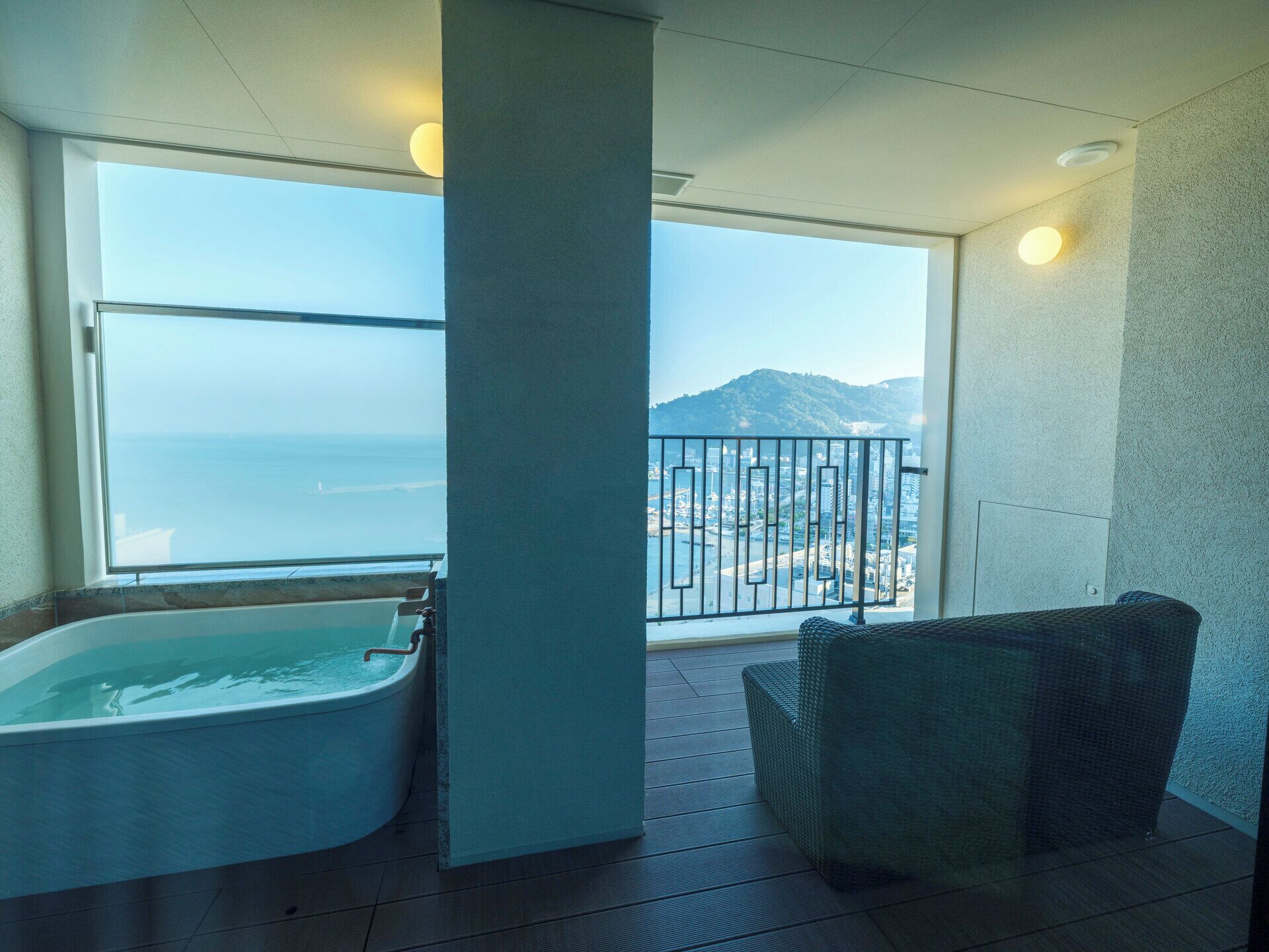 Un balcon avec vue sur la mer et les montagnes, une baignoire et un fauteuil en osier.