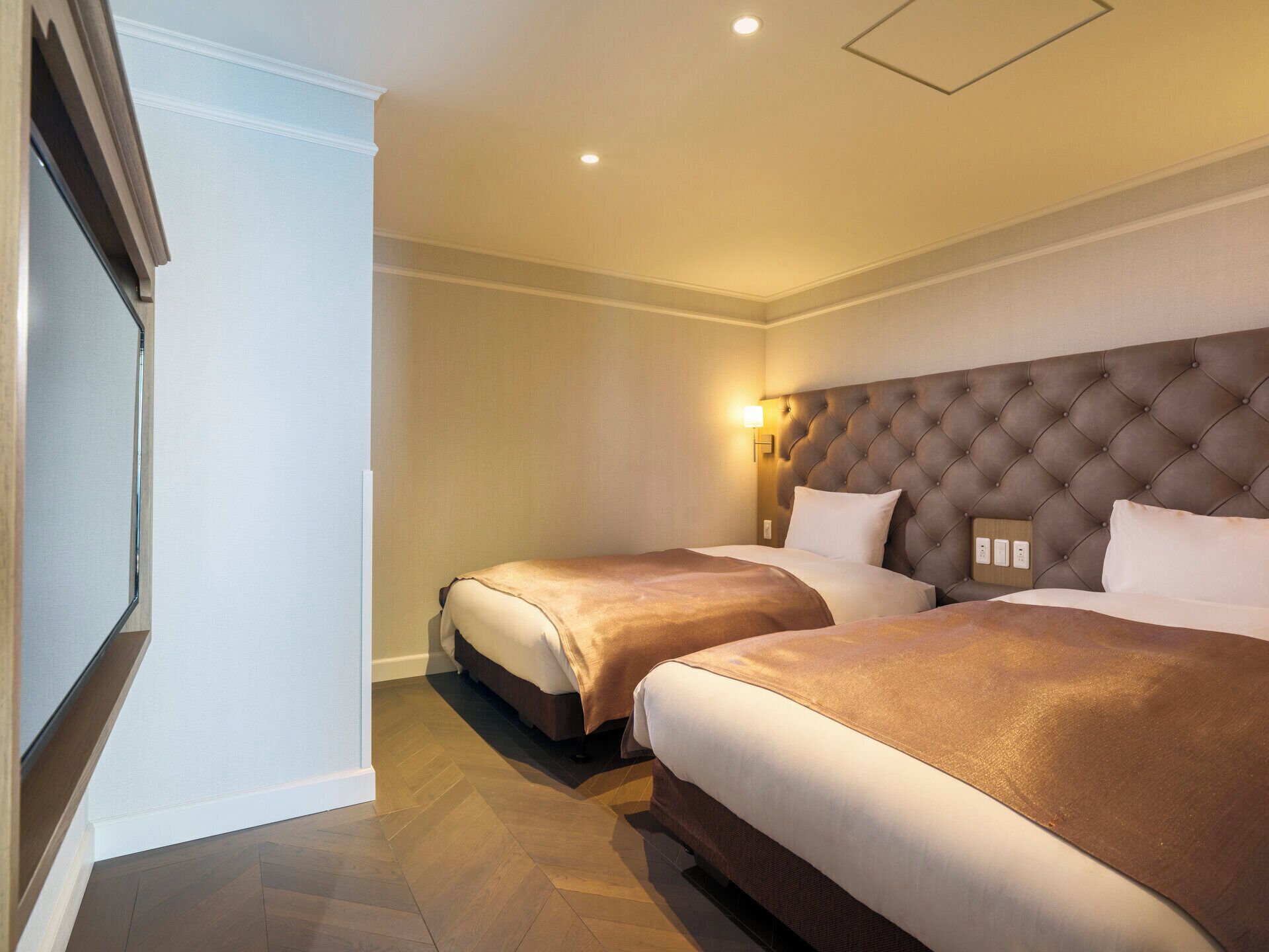 Ein Hotelzimmer mit zwei Betten, einem großen, rautenförmig geprägten Kopfteil, einer Wandleuchte und einem Holzboden.