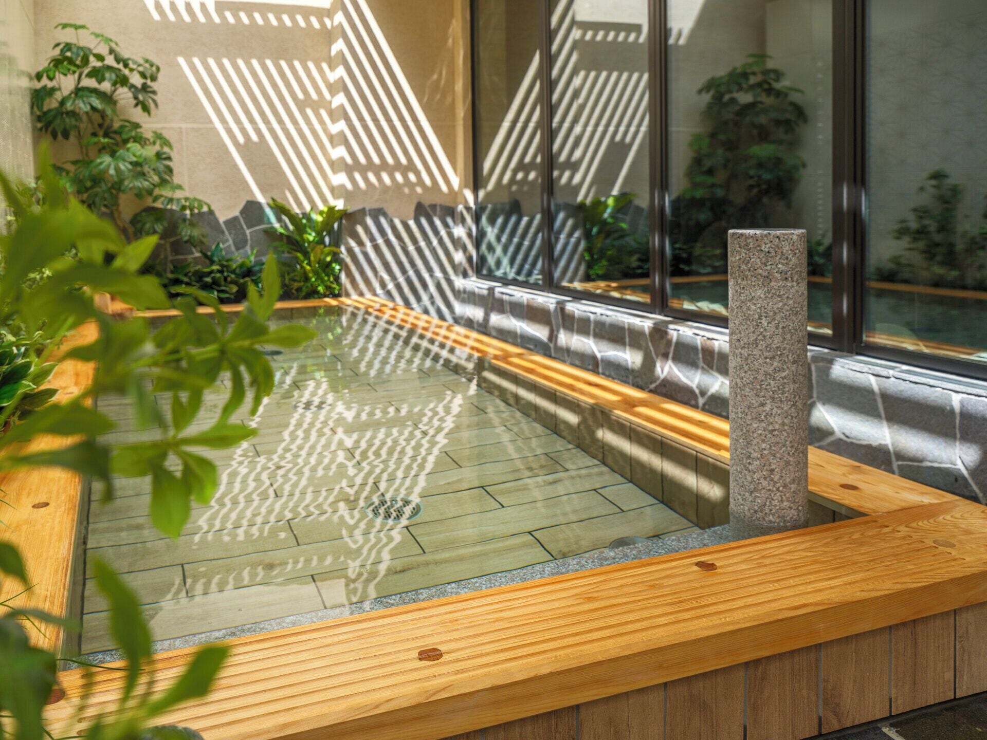 Un área de piscina interior moderna con cerramiento de vidrio, bancos de madera y abundante vegetación circundante.
