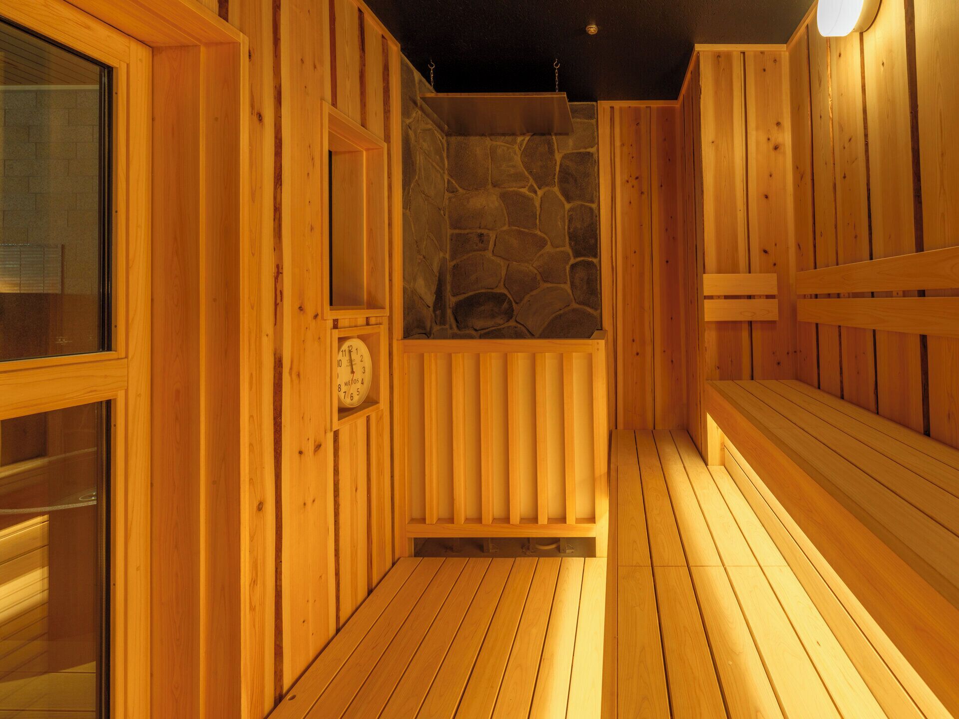 A wooden sauna with benches, a stone wall, and a clock.