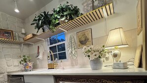 Una cocina con encimera blanca, un fregadero, un escurreplatos y una ventana. Hay plantas en la encimera y una lámpara en el mostrador.
