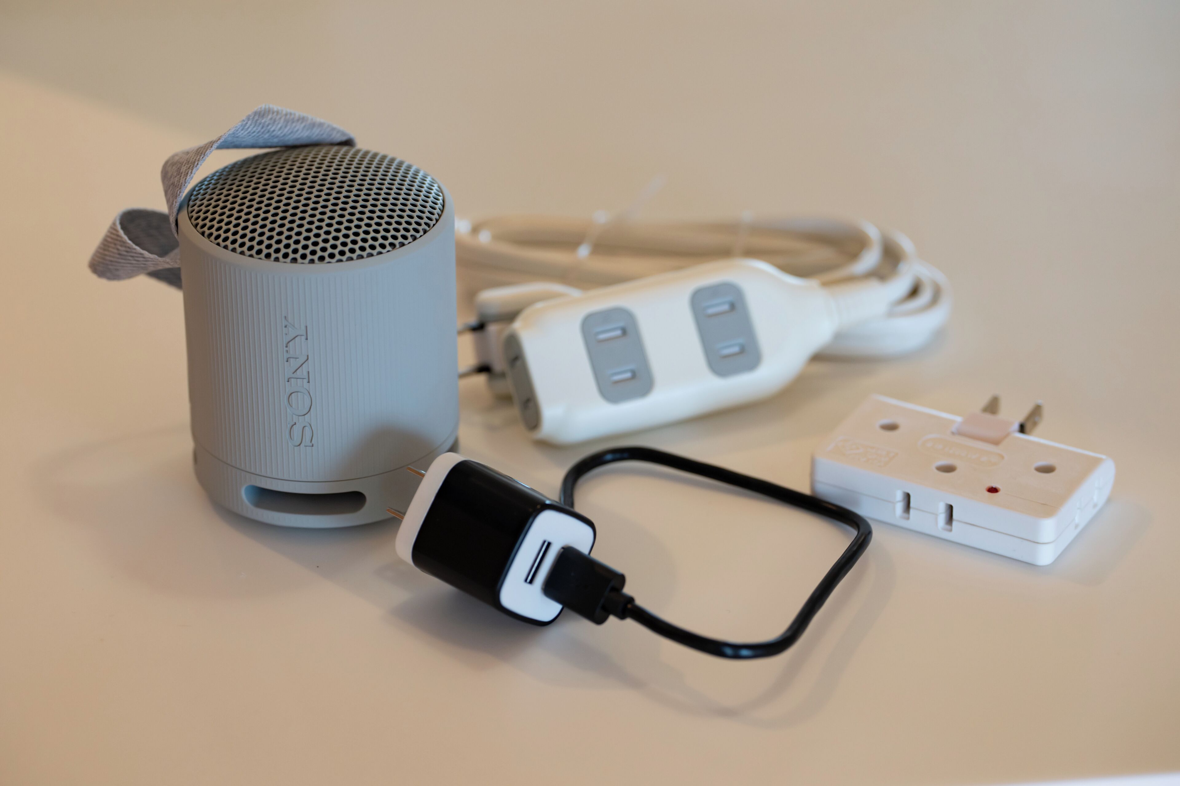 A grey cylindrical speaker with a fabric handle, a white power strip with two outlets, and a black power cord.