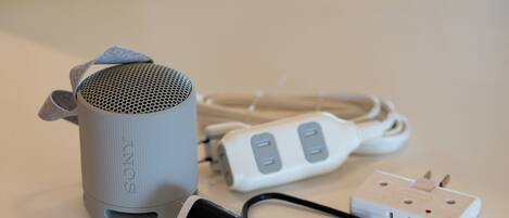 A grey cylindrical speaker with a fabric handle, a white power strip with two outlets, and a black power cord.