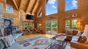 A cozy cabin interior with a stone fireplace, wooden walls, a large window, and a comfortable seating area. - Three Peaks Retreat: Pool, Hot Tub & Fireplace (Hyde Park)