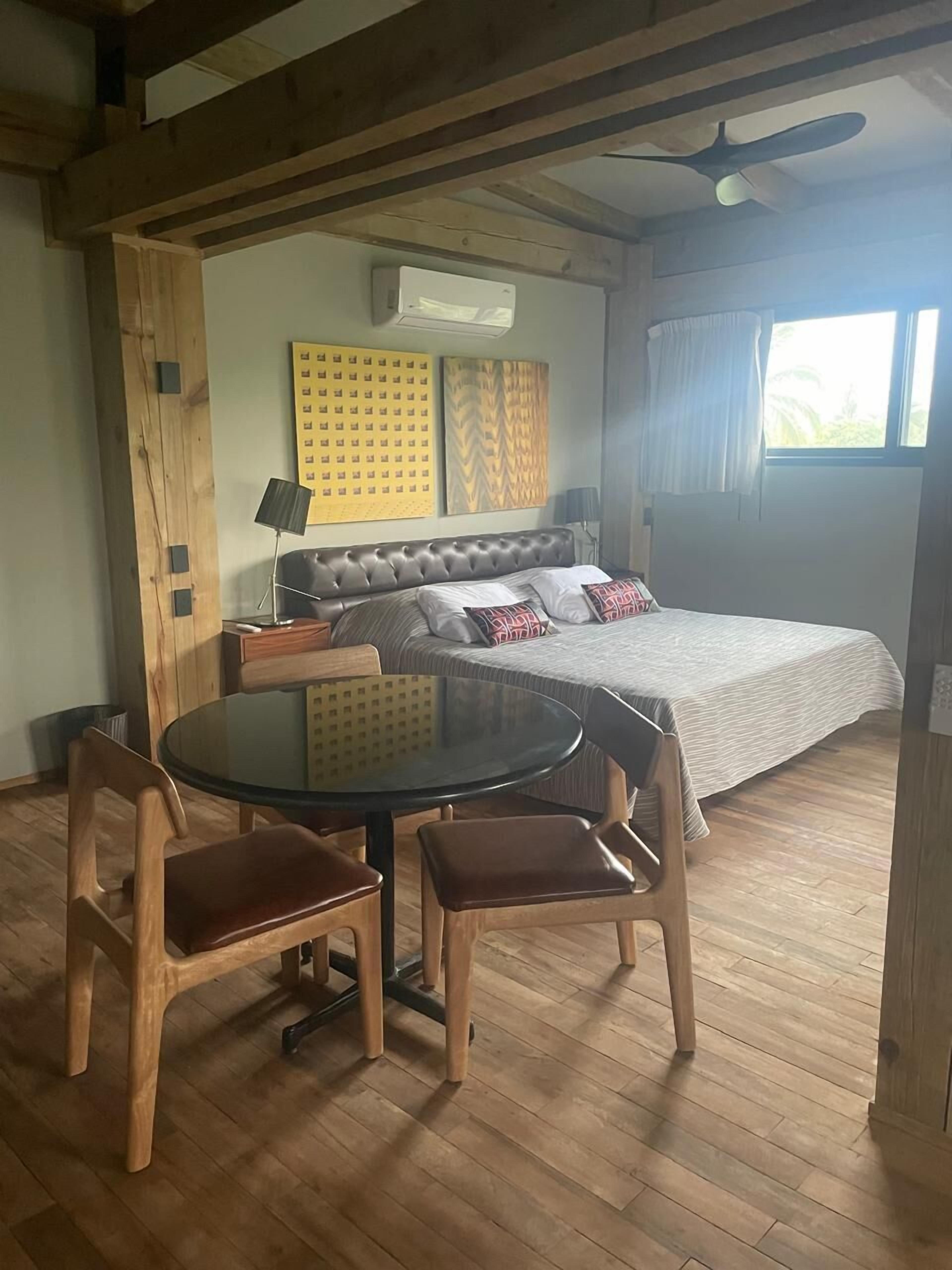 Una camera da letto con un letto, un tavolino rotondo in vetro e sedie, pavimento in legno e una ventola a soffitto.