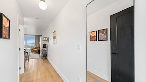 A hallway with wooden flooring, white walls, and a black door. There are framed pictures on the wall and a ceiling light. - Harbor Hub Retreat | Walk to Tours & Dining (Juneau)
