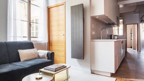A modern living room with a sofa, coffee table, and a bookshelf. A kitchen area with a sink and stove is visible in the background. - Blueground | Sainte-Avoye, view, nr Le Marais (Paris)