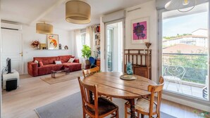 A modern living room with a red sofa, wooden dining table, and large windows offering a view of buildings and greenery. - LE CACTUS - Gorge de loup apartment (Lyon)
