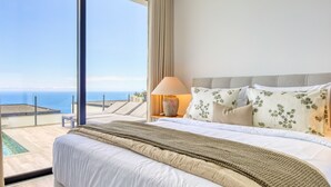 A bedroom with a large bed, a nightstand with a lamp, and a view of the ocean. - Villa Arco-Iris, a Home in Madeira (Arco da Calheta)