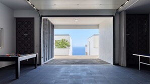 A modern indoor space with a pool table, a billiards table, and a view of the sea through an open door. - Villa Arco-Iris, a Home in Madeira (Arco da Calheta)