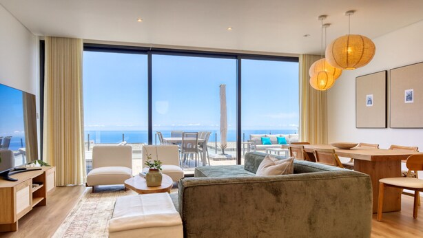 A modern living room with a large sofa, a dining table, and a view of the sea through large windows. - Villa Arco-Iris, a Home in Madeira (Arco da Calheta)
